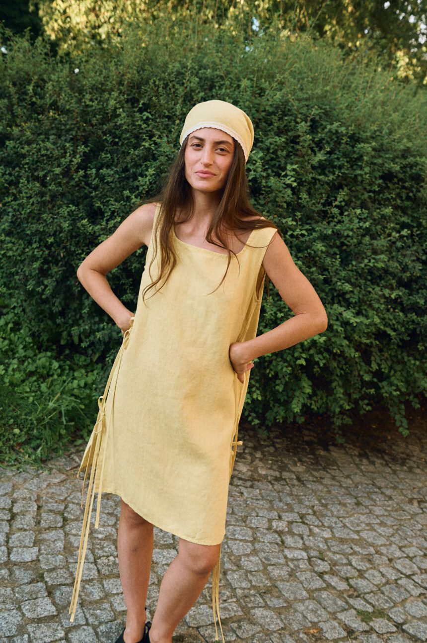 Model wearing a sleeveless butter yellow linen midi dress with a scoop neckline, side tie details, and a matching linen head wrap bandana