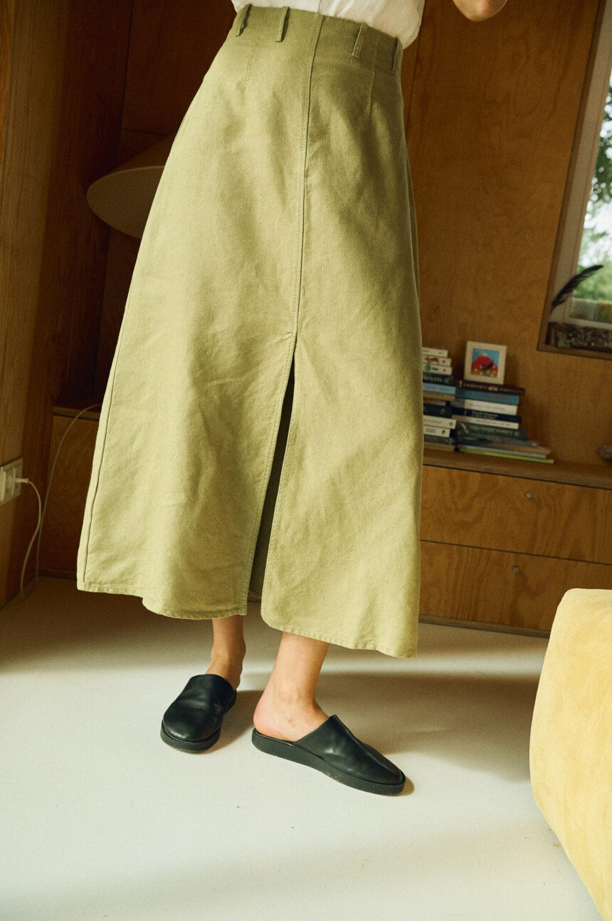 Close-up of a high-waisted olive green midi skirt made from heavy linen, featuring a front slit, belt loops, and a structured A-line cut