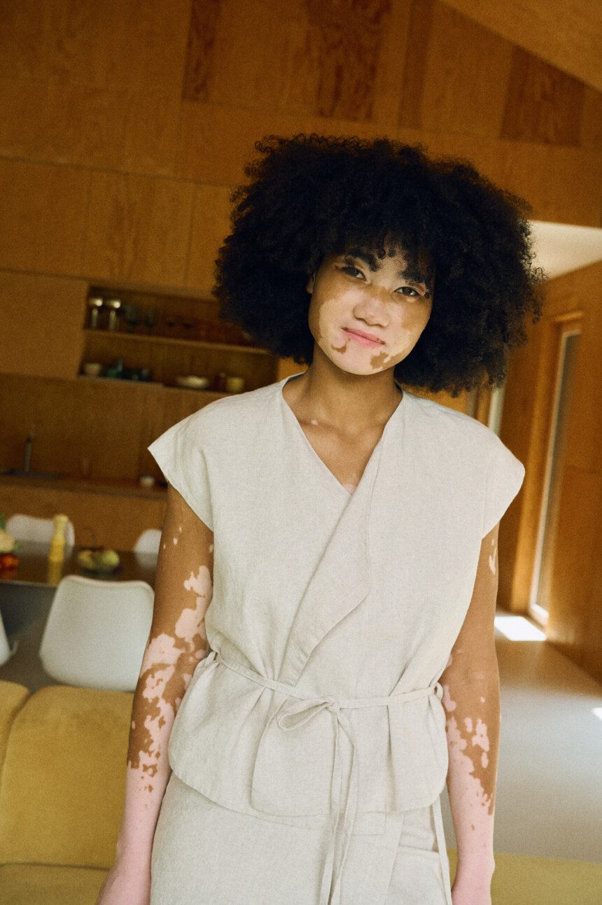 Woman wearing a white linen cotton wrap vest with tie belt standing in a modern living room.