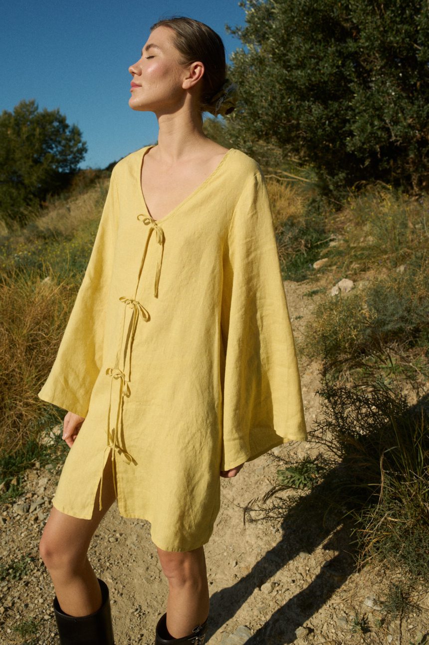 Model wearing a mini-length linen dress in butter color, featuring a deep V-neckline, four decorative front tie-bow strings, wide shoulders, and flowy sleeves, all in a relaxed A-line silhouette and oversized fit